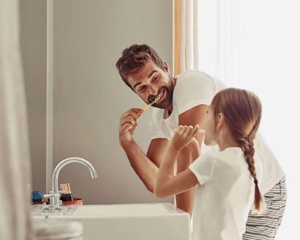 Father and daughter brushing their teeth together at a sink.