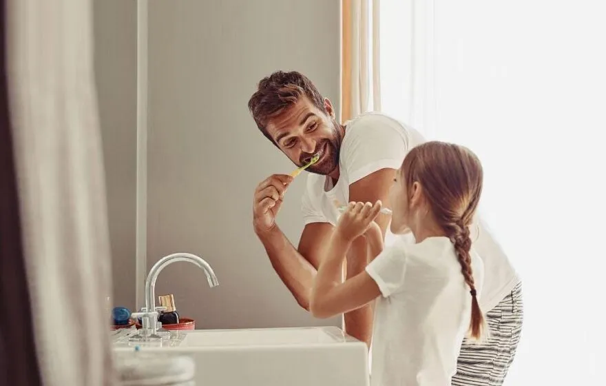 Father and daughter brushing their teeth together at a sink.